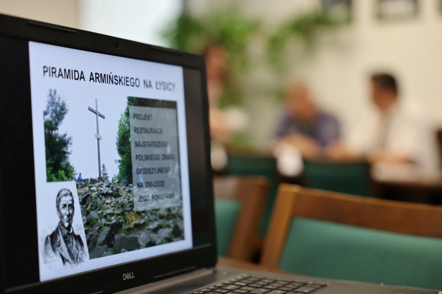 Na ekranie laptopa widnieje zdjęcie krzyża na szczycie góry z tekstem "Piramida Armińskiego na Łysicy". W tle siedzą ludzie.