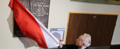 Mężczyzna z biało-czerwoną flagą przy tablicy pamiątkowej Jerzego Sozańskiego. Obok drzwi z numerem 1.18a.
