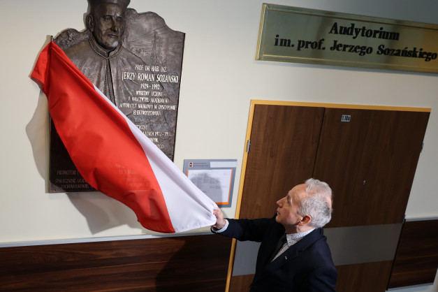 Mężczyzna z biało-czerwoną flagą przy tablicy pamiątkowej Jerzego Sozańskiego. Obok drzwi z numerem 1.18a.