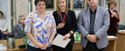 Trzy osoby stoją przed portretami. Kobieta po lewej ma na sobie kolorową bluzkę. Kobieta w środku trzyma medal i dokument.