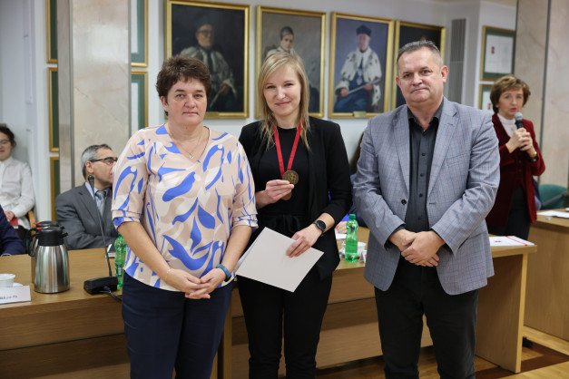 Trzy osoby stoją przed portretami. Kobieta po lewej ma na sobie kolorową bluzkę. Kobieta w środku trzyma medal i dokument.