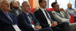 Grupa mężczyzn w garniturach siedzi na krzesłach, słuchając. Kontekst: konferencja lub wydarzenie biznesowe.