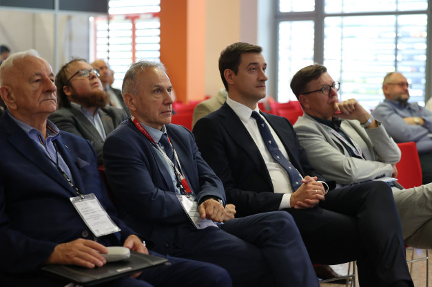 Grupa mężczyzn w garniturach siedzi na krzesłach, słuchając. Kontekst: konferencja lub wydarzenie biznesowe.