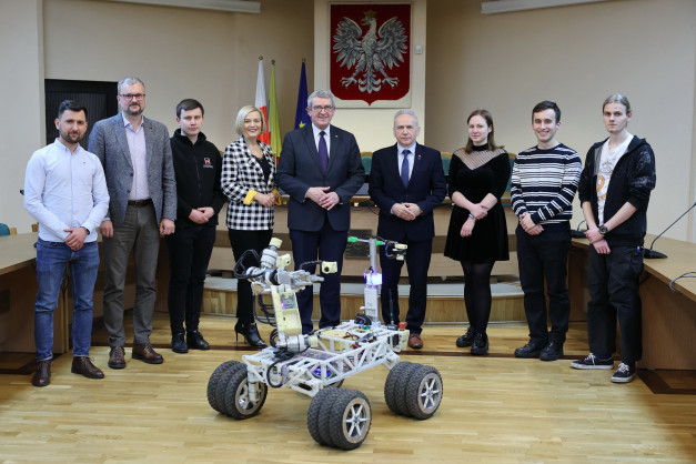 Grupa osób pozuje obok łazika marsjańskiego. W tle godło Polski i flagi.