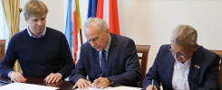 Trzech mężczyzn podpisuje dokumenty przy stole. W tle flagi Polski i Unii Europejskiej.