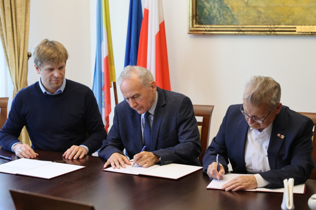 Trzech mężczyzn podpisuje dokumenty przy stole. W tle flagi Polski i Unii Europejskiej.