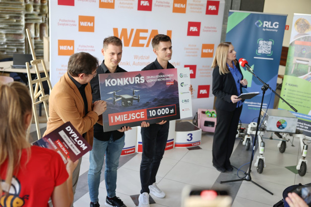 Dwóch mężczyzn trzyma tablicę z napisem "I MIEJSCE 10 000 zł". Kontekst: wydarzenie związane z konkursem.