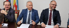 Trzech mężczyzn w garniturach siedzi przy stole. Flagi i obraz wiszą na ścianie. Kontekst: spotkanie.
