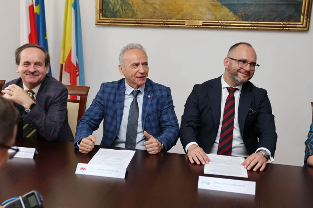 Trzech mężczyzn w garniturach siedzi przy stole. Flagi i obraz wiszą na ścianie. Kontekst: spotkanie.