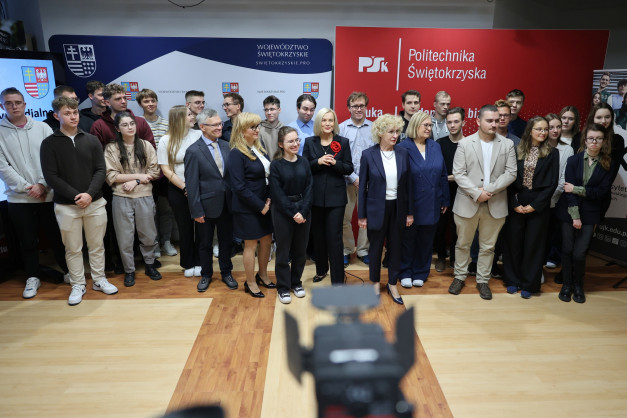 Grupa osób stoi przed banerami z logo województwa i Politechniki Świętokrzyskiej. Kluczowe obiekty: ludzie, banery, logo.