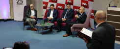 Panel dyskusyjny z udziałem czterech mężczyzn w garniturach, siedzących na krzesłach.