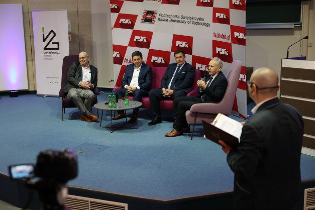 Panel dyskusyjny z udziałem czterech mężczyzn w garniturach, siedzących na krzesłach.