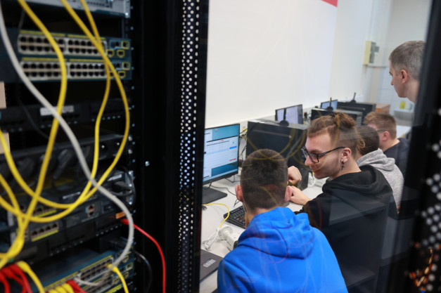 Grupa osób pracuje przy komputerach w serwerowni. Widoczne są serwery, kable sieciowe i monitory.