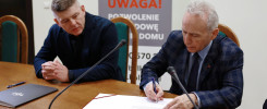 Dwóch mężczyzn przy stole podpisuje dokumenty. Na stole znajdują się mikrofony i teczka.