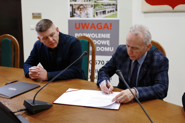 Dwóch mężczyzn przy stole podpisuje dokumenty. Na stole znajdują się mikrofony i teczka.