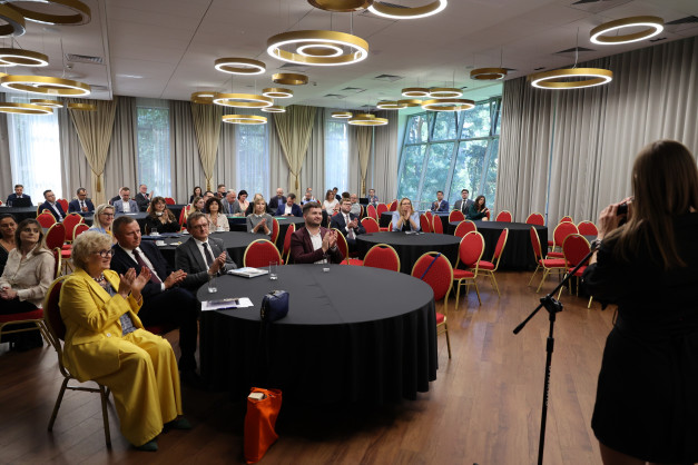 Ludzie siedzą przy okrągłych stołach w sali konferencyjnej. W tle okna z widokiem na drzewa.