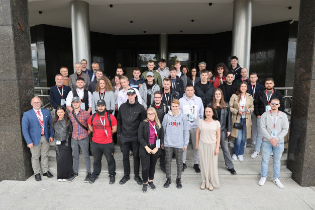 Grupa ludzi pozuje do zdjęcia przed budynkiem. Kluczowe obiekty: ludzie, budynek, schody. Kontekst: wydarzenie grupowe.
