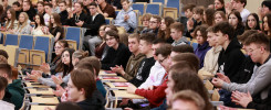 Grupa młodych ludzi siedzi w sali wykładowej, wielu z nich klaszcze. Kluczowe obiekty: ludzie, krzesła, stoły.