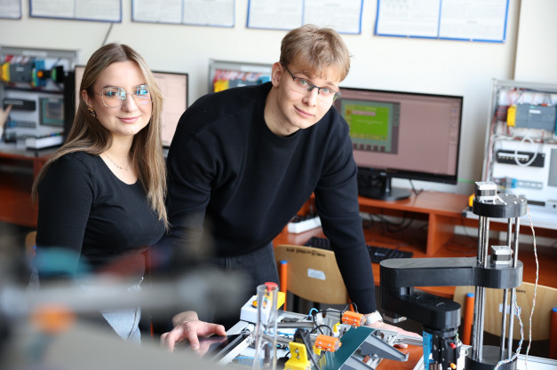 Dwoje studentów w okularach pracuje przy robocie. W tle komputery i sprzęt laboratoryjny.