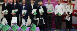 Osoby w garniturach i sukience stoją na scenie, trzymając dyplomy i torby. W tle banery.
