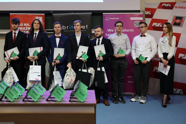 Osoby w garniturach i sukience stoją na scenie, trzymając dyplomy i torby. W tle banery.