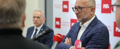 Mężczyzna w okularach i garniturze udziela wywiadu przed mikrofonami. Tło to ściana z logo PŚK.