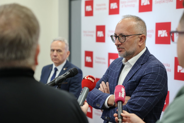Mężczyzna w okularach i garniturze udziela wywiadu przed mikrofonami. Tło to ściana z logo PŚK.