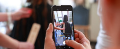 Osoba trzyma telefon, którym fotografuje fryzjera mierzącego włosy klientki linijką.
