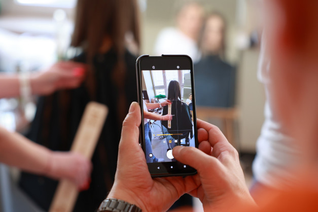 Osoba trzyma telefon, którym fotografuje fryzjera mierzącego włosy klientki linijką.