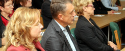 Ludzie siedzą przy stołach, słuchając. Widać kobiety i mężczyzn w formalnych strojach.