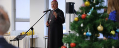 Biskup w stroju duchownym przemawia przy mikrofonie. W tle choinka i stół z napojami.