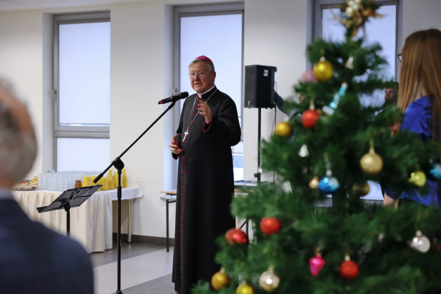 Biskup w stroju duchownym przemawia przy mikrofonie. W tle choinka i stół z napojami.
