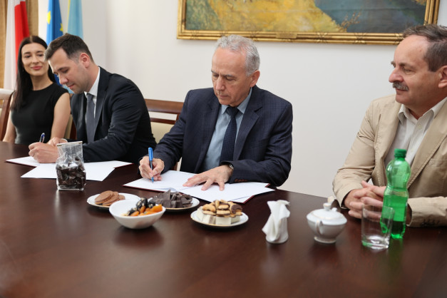 Ludzie przy stole podpisują dokumenty. Na stole są słodycze, napoje i flagi.