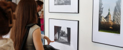 Wystawa fotografii. Kobieta ogląda czarno-białe zdjęcie z arkadami i kolorowe zdjęcie z wieżą kościelną.