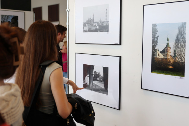 Wystawa fotografii. Kobieta ogląda czarno-białe zdjęcie z arkadami i kolorowe zdjęcie z wieżą kościelną.
