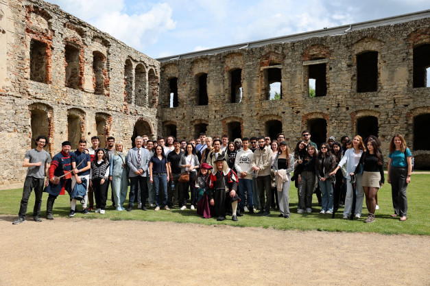 Grupa ludzi pozuje do zdjęcia na tle ruin zamku. Wśród nich są osoby w strojach historycznych.