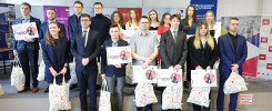 Grupa osób pozuje do zdjęcia z czekami na 1000 zł i torbami. Kluczowe obiekty: ludzie, czeki, torby.