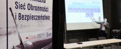 Biały baner z napisem "Politechniczna Sieć Obronnosci i Bezpieczeństwa" oraz fragment broni. W tle ekran projekcyjny.