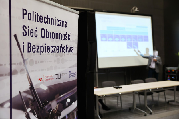 Biały baner z napisem "Politechniczna Sieć Obronnosci i Bezpieczeństwa" oraz fragment broni. W tle ekran projekcyjny.