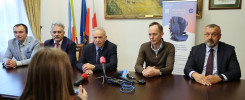 Pięciu mężczyzn siedzi przy stole, przed nimi mikrofony i baner. Kontekst: konferencja prasowa.