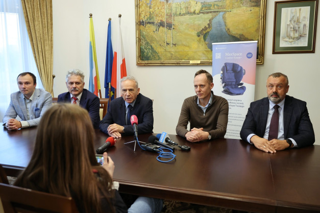 Pięciu mężczyzn siedzi przy stole, przed nimi mikrofony i baner. Kontekst: konferencja prasowa.