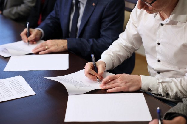 Ludzie podpisują dokumenty na ciemnym stole. Kluczowe obiekty: dokumenty, długopisy, dłonie.
