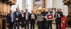 Grupa osób w kościele pozuje do zdjęcia z nagrodami. W tle obraz religijny i drewniane ławy.
