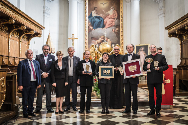 Grupa osób w kościele pozuje do zdjęcia z nagrodami. W tle obraz religijny i drewniane ławy.