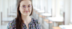 Młoda kobieta w okularach i kwiecistej bluzce stoi w jasnym, przestronnym korytarzu z rzędami stołów.
