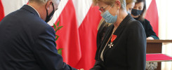 Mężczyzna w garniturze podaje gałązkę kobiecie w czarnej marynarce z orderem. W tle polskie flagi.