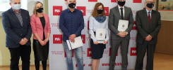 Osoby w maseczkach stoją przed banerem Politechniki Świętokrzyskiej. Kluczowe obiekty: ludzie, baner, dokumenty.