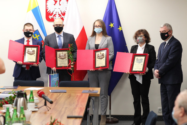 Osoby w maseczkach odbierają nagrody. W tle flagi Polski i UE. Kluczowe obiekty: ludzie, nagrody, flagi.