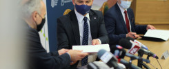 Trzech mężczyzn w garniturach, dwóch w maseczkach, siedzi przy stole z mikrofonami. Jeden podaje dokument.