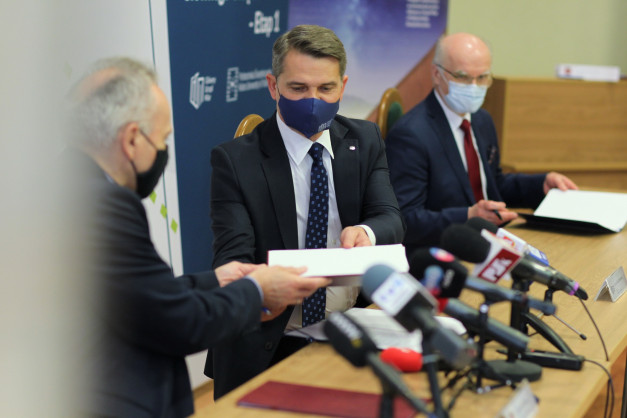 Trzech mężczyzn w garniturach, dwóch w maseczkach, siedzi przy stole z mikrofonami. Jeden podaje dokument.